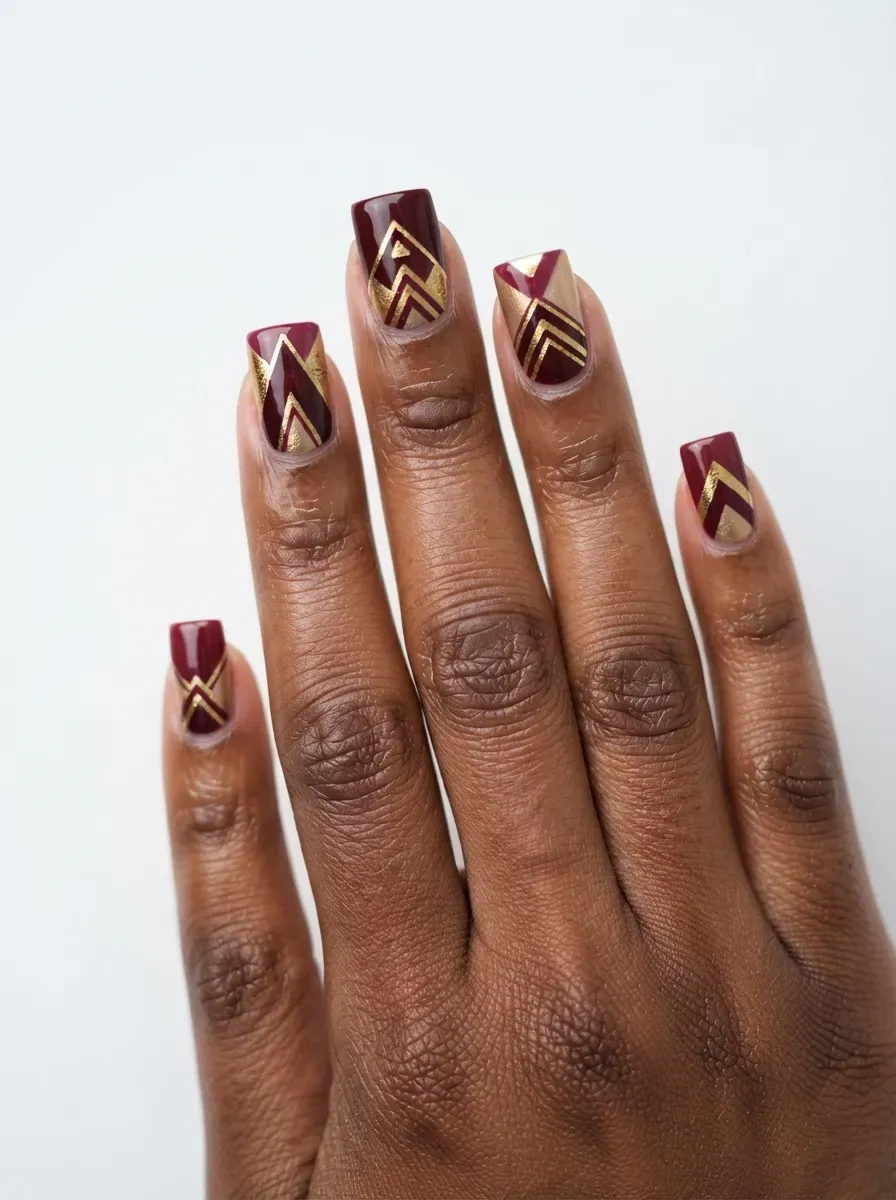 Close-up of medium square nails with burgundy and gold geometric design on deep chestnut skin