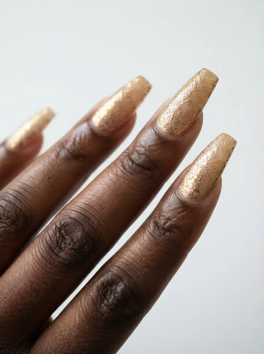 Close-up of coffin nails with gold foil lace over nude polish on rich dark skin