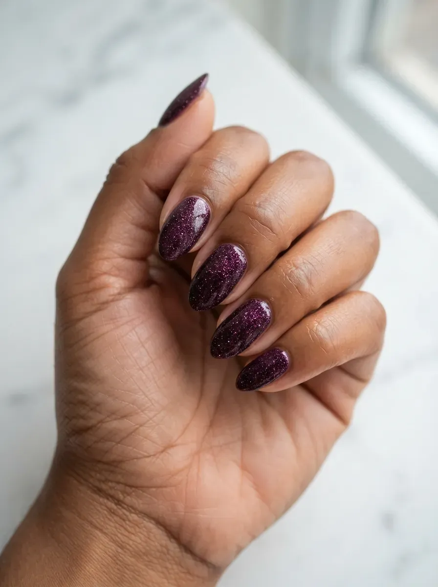 Close-up of medium almond nails with midnight plum glitter polish on deep brown skin